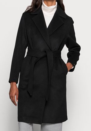 Czarny wełniany płaszcz z paskiem, szerokimi klapami, noszony na białym golfie i jasnoszarych spodniach, ręka w kieszeni, fason o długości do połowy uda.