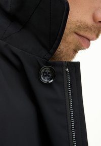 Schwarze Jacke mit hohem Kragen, glänzendem Knopf-Akzent und sichtbarem Reißverschluss. Das Material wirkt glatt mit diagonalen Nähdetails.