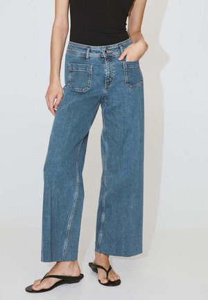 Femme portant un jean taille haute, jambes larges, bleu avec de grandes poches avant et un haut noir sans manches, debout avec une main dans la poche.