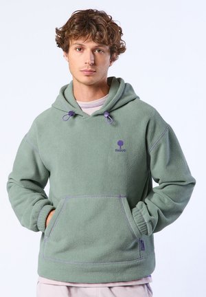 Jeune homme aux cheveux bouclés portant un sweat à capuche en polaire vert sauge avec poche frontale et détails de cordon violet, debout devant un fond uni.