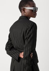 Svart blazer med pinstriper og belte i midjen, med skreddersydd passform, strukturerte skuldre og en jevn tekstur. Grå solbriller og gull detaljer.