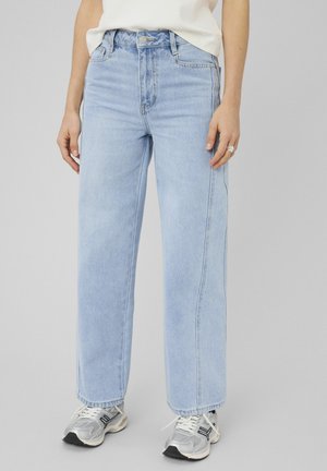 Jean bleu clair taille haute à jambes droites, porté avec des baskets blanches et grises et un haut blanc rentré, mains détendues le long du corps.