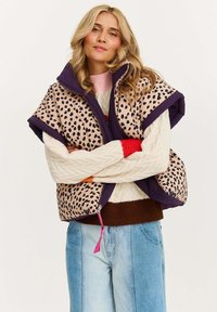 Kamizelka puchowa w panterkę na beżowym tle, fioletowe wykończenie i kieszeń na zamek. Noszona na kremowym swetrze i niebieskich dżinsach.