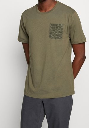 Homme portant un t-shirt olive à manches courtes avec une poche à motif et un pantalon gris foncé, debout devant un fond uni.