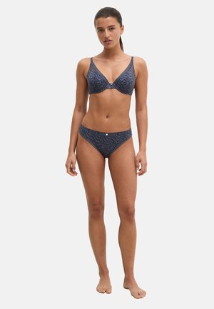 Mujer de pie descalza con conjunto a juego de sujetador y ropa interior azul oscuro con estampado sobre fondo claro liso.