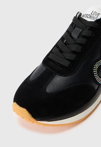Svart sneaker med suede- og skinnmateriale, gummisåle, rund tå og rhinstone-logo detalj på siden.