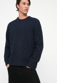 Tmavo modrý pletený sveter s textúrovaným geometrickým vzorom, okrúhlym výstrihom a rebrovanými manžetami. Skombinované s čiernymi nohavicami.
