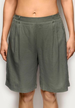 Osoba nosząca luźne, oliwkowozielone krótkie spodenki do kolan z przednimi zakładkami i bocznymi kieszeniami, stojąca na tle jednolitego tła.