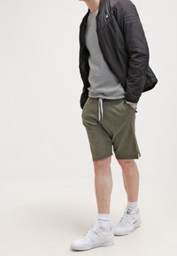 Zwarte bomberjack, grijs textuur trui met ronde hals, olijfgroene shorts met trekkoord, witte sneakers en witte sokken. Minimalistische sportieve outfit.