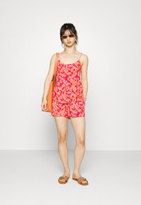 Macacão vermelho com padrão floral, apresentando alças finas, bolsos e um corte relaxado; combinado com sandálias castanhas e uma bolsa tote laranja.
