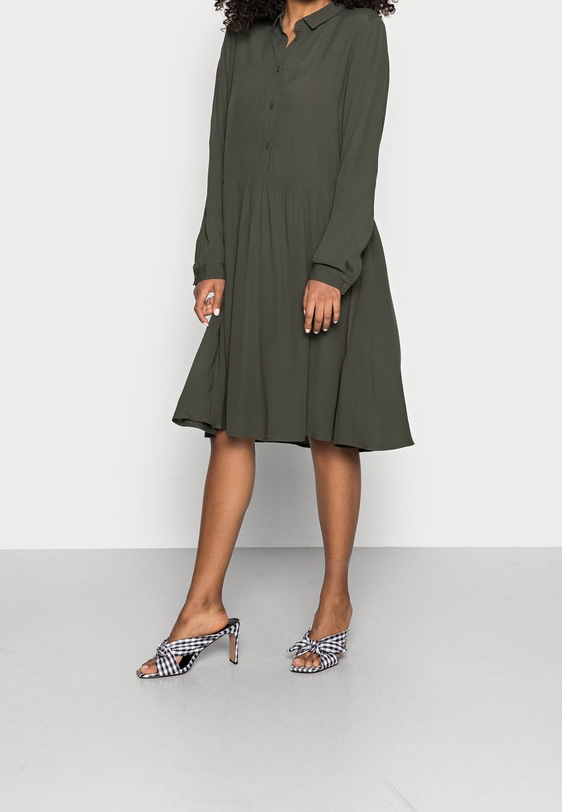 Kobieta w długim rękawie, ciemnooliwkowej sukience midi, połączona z czarno-białymi, w kratkę, wysokimi sandałami w stylu slip-on.