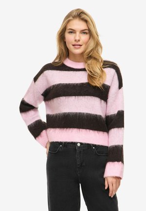 Pitkähiuksinen vaaleatukkainen nainen, joka pukeutuu vaaleanpunaiseen ja tummanruskeaan raidalliseen pörröiseen neulepaitaan ja mustiin farkkuihin, seisoo yhdellä kädellä lanteilla.