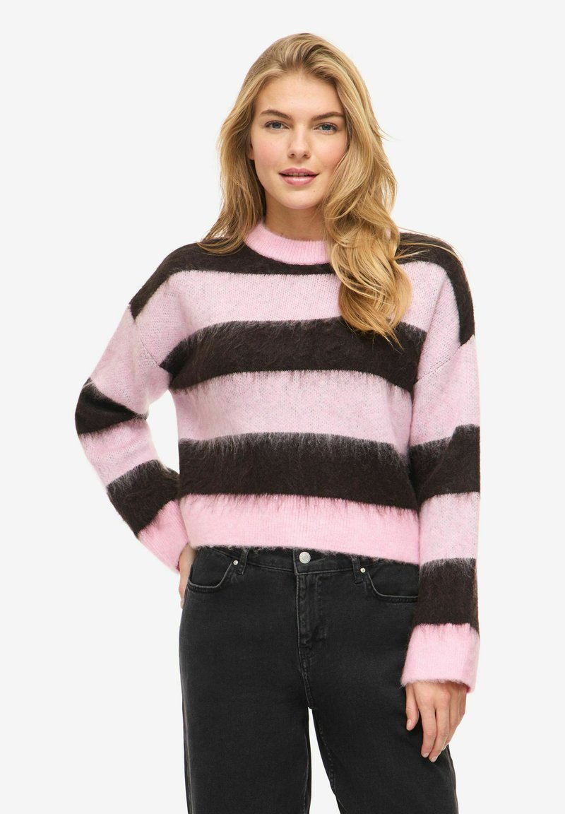 Femme aux longs cheveux blonds portant un pull duveteux rayé rose et marron foncé, ainsi qu'un jean noir, debout avec une main sur la hanche.