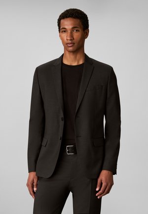 Homme portant une veste de costume noire, une chemise noire et un pantalon noir, debout devant un fond gris uni.