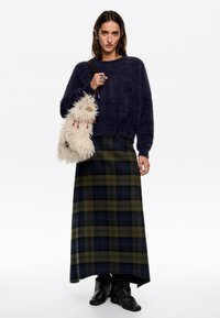 Granatowy futrzany sweter, długa zielono-granatowa spódnica w kratę, czarne botki za kostkę. Trzyma puszystą białą torbę z czerwonymi akcentami i dekoracyjnym okuciem.