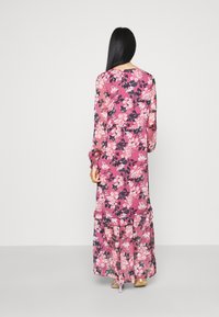 Kvinna med långt svart hår iklädd en lång rosa blommig klänning och lila högklackade sandaler, stående vänd bortåt mot en enfärgad bakgrund.