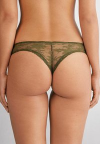 Olijfgroene kantstring met doorschijnende stof, bloemmotief en elastische tailleband; heeft kleine openingen voor een minimalistisch dekkend ontwerp.