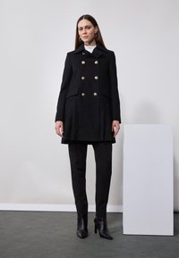 Manteau noir à double boutonnage avec des boutons dorés, poches latérales et coupe ajustée. Porté sur un pantalon noir et des bottes à talons hauts et pointues.