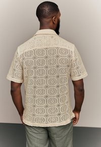 Beżowa koszula z krótkim rękawem z wzorem szydełkowym zawierającym ośmiokątne motywy. Lekki, przejrzysty materiał o teksturowanym wykończeniu.