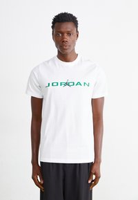 HVIT bomulls T-skjorte med grønn "JORDAN" tekst og svart Jumpman-logo. Klassisk rund hals og korte ermer. Glatt tekstur.