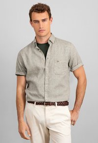 Cămașă cu mânecă scurtă, cu nasturi, de culoare gri deschis, cu un buzunar pe piept, purtată cu un tricou verde închis și pantaloni bej, strânși cu o curea împletită de culoare brună.