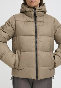 Beige puffer-jakk, millel on kapuuts, ees lukuga ja küljetaskudega. Materjal on siledast tekstuurist ja vatitud polsterdusega.
