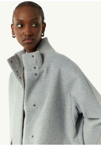 Graue Wollmischungjacke mit hohem, geripptem Kragen und Druckknopfverschluss. Bemerkenswerte Details sind die glatte Textur und die minimalistischen Designelemente.
