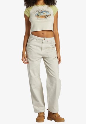 Donna con maglietta corta grafica verde chiaro e beige, pantaloni beige a vita alta e stivali marrone chiaro con lacci, in piedi con le braccia rilassate.