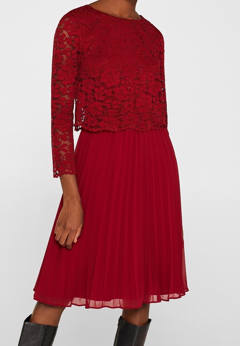 Femme portant un top en dentelle rouge à manches longues et une jupe midi plissée associée à des bottes noires montantes.