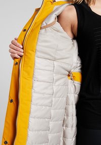 Veste extérieure jaune avec une texture brillante, dotée d'une doublure intérieure matelassée beige. Le design comprend une fermeture à glissière avant et des boutons-pression.