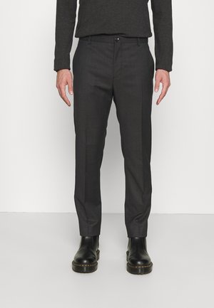 Homme portant un pantalon de costume gris foncé ajusté et des bottes en cuir noires, debout devant un fond clair uni.
