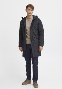 Mørk grå lang vinterjakke med hette, med glidelås og trykknapper, kombinert med en beige genser og mørke jeans.