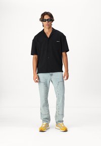 Chemise noire à manches courtes avec deux poches à l'avant, jeans en denim bleu clair et baskets jaunes. Comprend des lunettes de soleil rectangulaires.