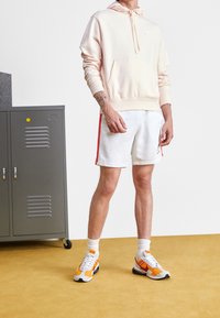 Ljusrosa hoodie med en framficka, vita shorts med röda sidostråk och flerfärgade sneakers. Grå skåp i bakgrunden.