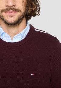 Bordó pulóver texturált kötött anyagból, kerek nyakkivágással és vállrész kiemelésekkel. Alatta egy világos kék kockás ing látható.