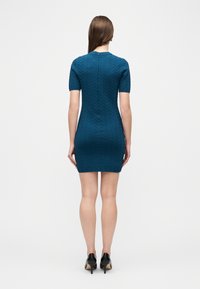 Γυναίκα που στέκεται γυρισμένη πλάτη, ντυμένη με εφαρμοστό κοντομάνικο φόρεμα σε τιρκουάζ απόχρωση με διακριτικό σχέδιο σε σχήμα ζιγκ-ζαγκ και μαύρα ψηλοτάκουνα παπούτσια.