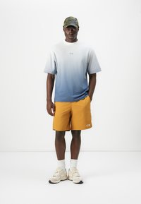 Light blue gradient t-shirt, orange shorts, and beige sneakers. Includes a camouflage cap and white socks. Features logo accents on shirt and shorts.