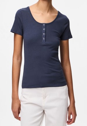 Vrouw draagt een marineblauw geribd Henley-T-shirt met korte mouwen en vier knopen en een witte broek, staand tegen een effen lichte achtergrond.