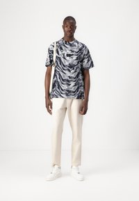 Short-sleeve t-shirt in navy and white wave pattern, featuring a round neckline and relaxed fit. Paired with light-colored trousers and white sneakers.