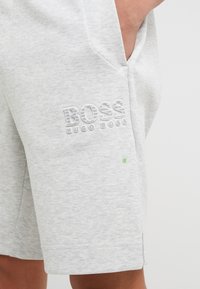 Shorts gris clair avec poche latérale et logo "BOSS HUGO BOSS" en relief sur la cuisse, portés par une personne avec la main dans la poche.