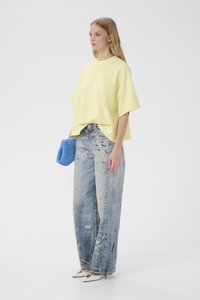 Ung kvinde iført løs gul top, brede blå slidte jeans, holder blå clutch og hvide spidse sko, stående på hvid baggrund.