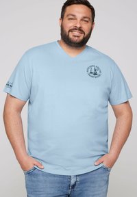 Camiseta de algodón azul claro con escote en V, mangas cortas y un logotipo gráfico redondo en el pecho, además de texto en la manga izquierda.