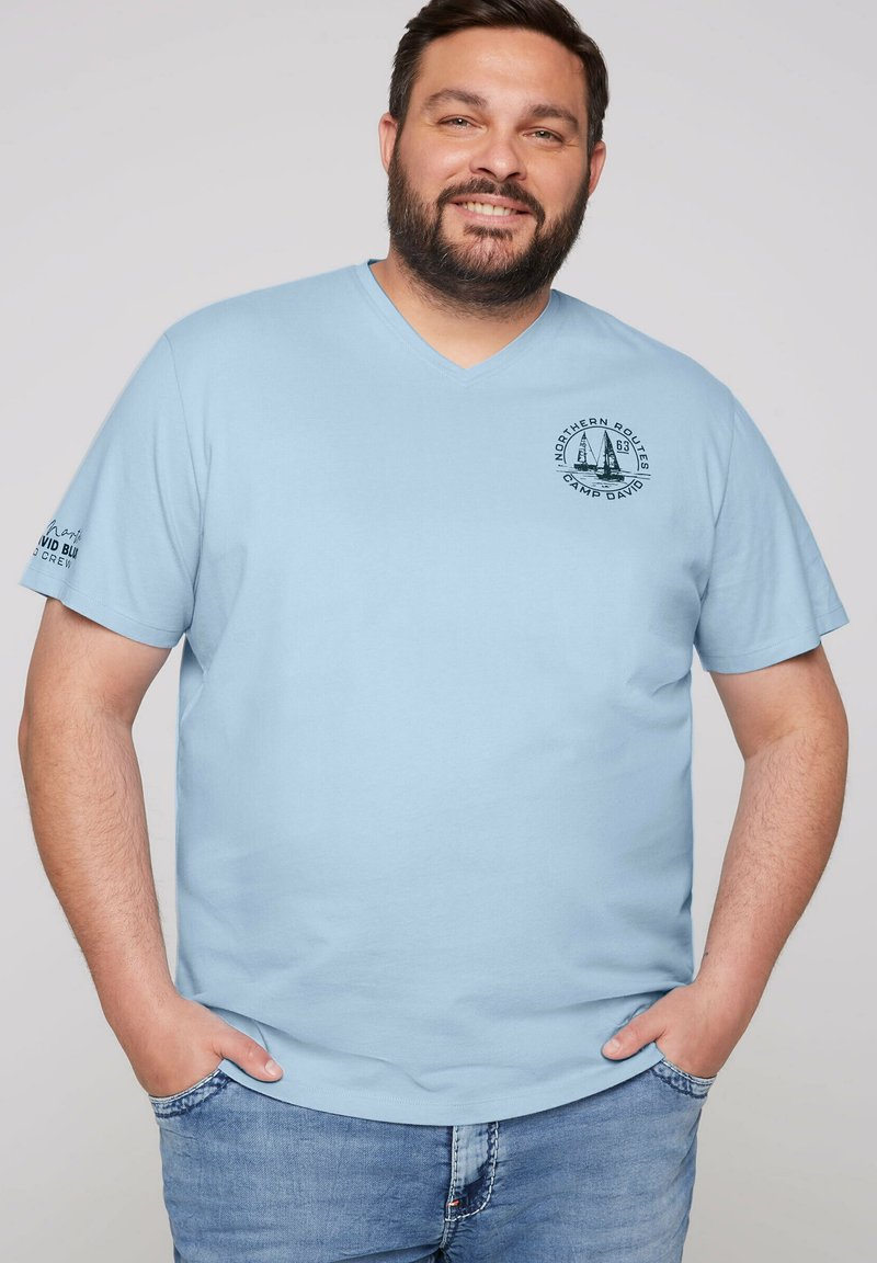 Camiseta de algodón azul claro con escote en V, mangas cortas y un logotipo gráfico redondo en el pecho, además de texto en la manga izquierda.