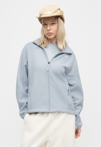 Ξανθός/ή φοράει ανοιχτό μπλε φλις ζακέτα με φερμουάρ, μπεζ γούνινο καπέλο με το λογότυπο The North Face και άσπρο παντελόνι, στέκεται μπροστά σε απλό φόντο.