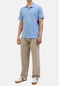 Hellblaues Poloshirt mit strukturiertem Muster, kombiniert mit beigen, gefalteten Hosen und weißen Turnschuhen mit blauen und braunen Akzenten.