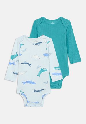 Δύο φορμάκια μωρού με μακρύ μανίκι, το ένα μονόχρωμο σε τιρκουάζ ριμπ, το άλλο λευκό με μοτίβα φαλαινών σε μπλε και τιρκουάζ, απλωμένα σε λευκό φόντο.