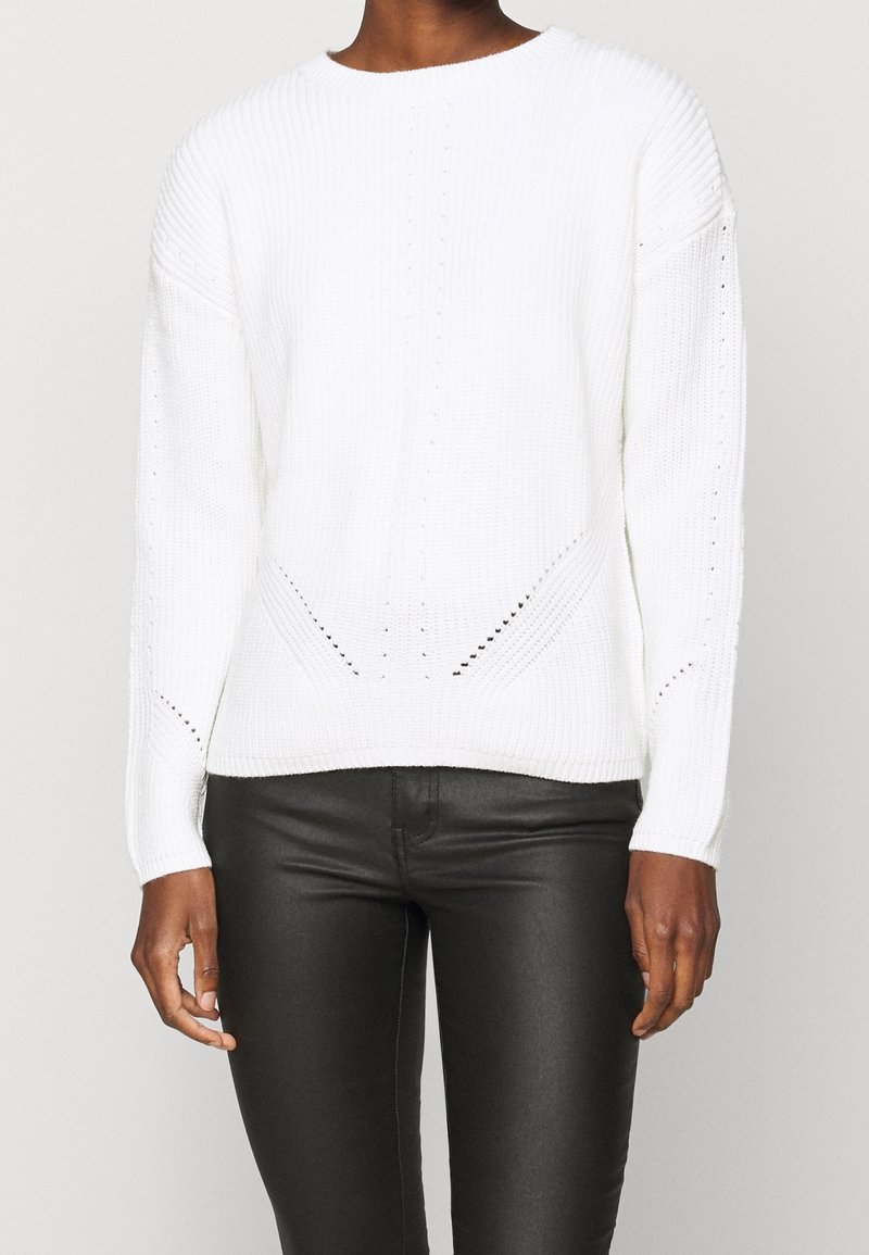 Persoon in een witte gebreide sweater met lange mouwen en subtiele patroondetails, gecombineerd met zwarte strakke broek voor een effen achtergrond.