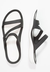 Sandales en caoutchouc noir avec une semelle blanche rembourrée, dotées de plusieurs sangles croisées pour un maintien sécurisé et d'une semelle intérieure texturée pour le confort.