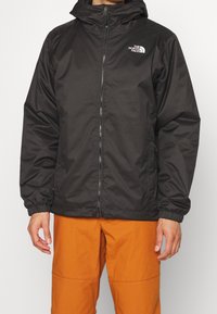 Giacca nera impermeabile con zip frontale, cappuccio regolabile, polsini elasticizzati e logo North Face sul petto sinistro. Indossata con pantaloni arancioni.