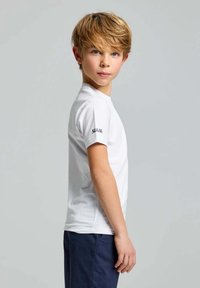 Weißes T-Shirt mit kurzen Ärmeln, runder Ausschnitt, sichtbare Stofftextur, mit schwarzem Logo auf dem linken Ärmel. Das Model trägt marineblaue Shorts.